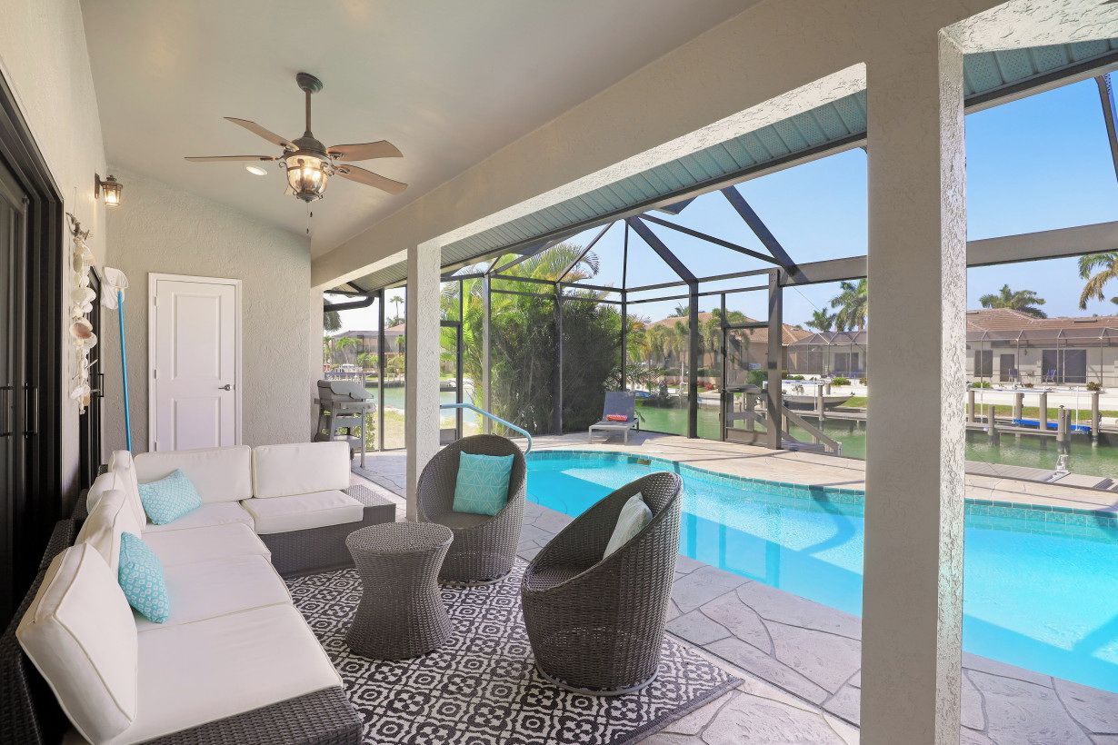 A covered outdoor area with a pool, white sofas, wicker chairs, and a ceiling fan.