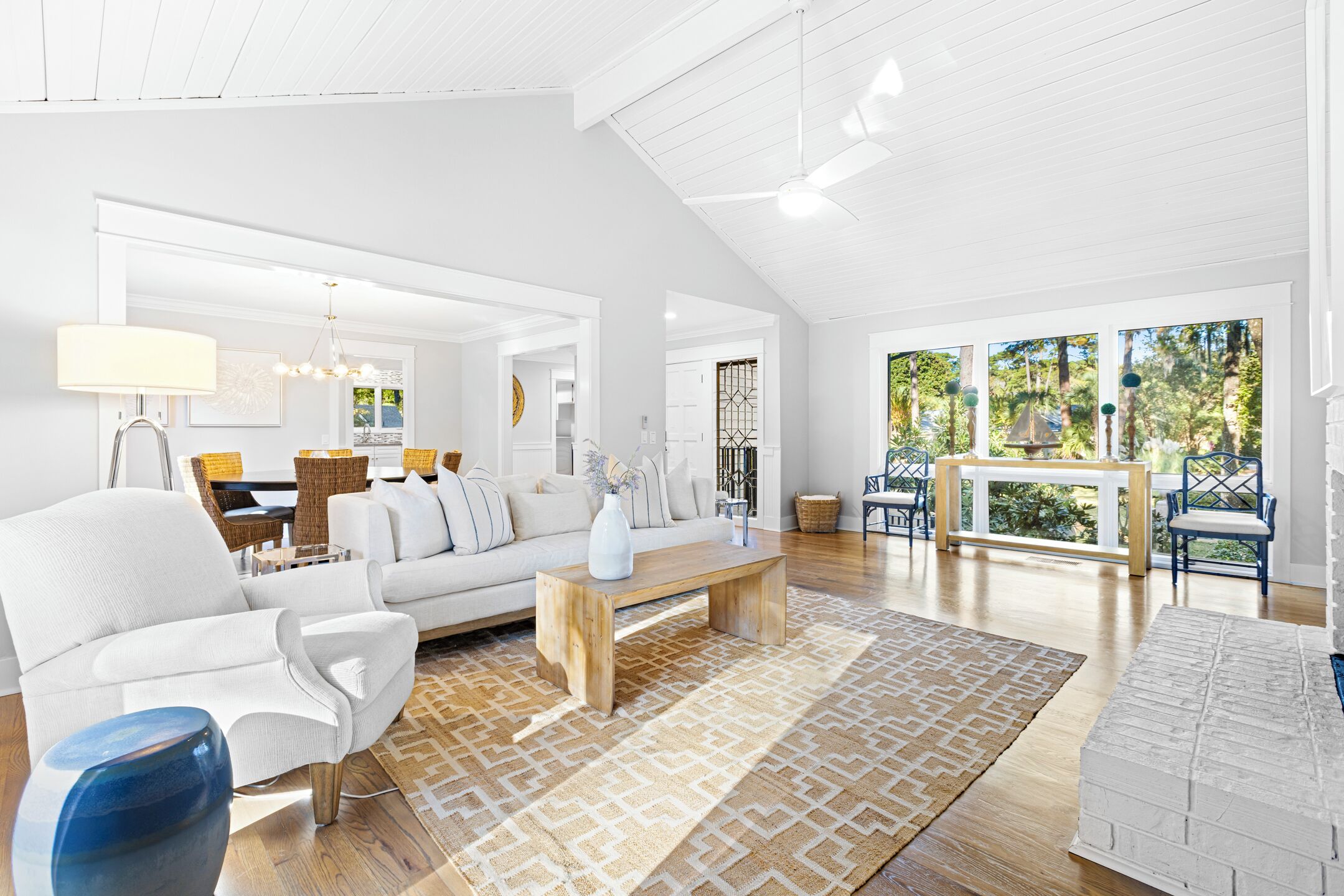 A spacious living room with a white sofa, a wooden coffee table, and a patterned rug. There is a dining area with a table and chairs, and a view of greenery outside.