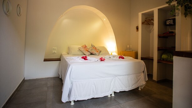 A neatly made bed with white bedding and red flowers, a bedside table with a lamp, and a small plant on the right. - Pousada Vila Nakau (Fernando de Noronha)