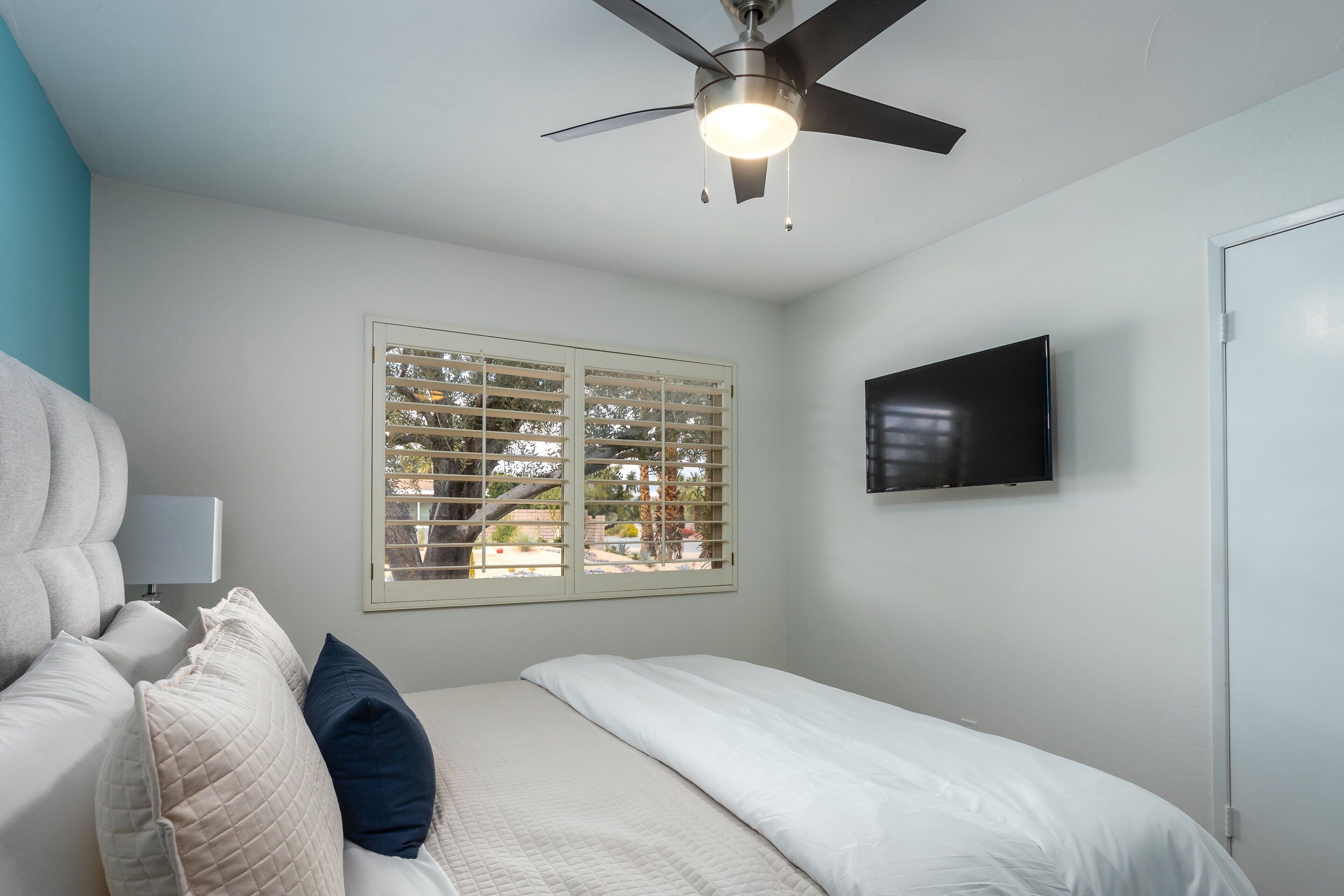 A bedroom with a bed, a ceiling fan, a TV, and a window with blinds.