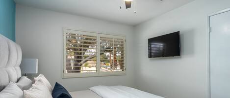A bedroom with a bed, a ceiling fan, a TV, and a window with blinds.