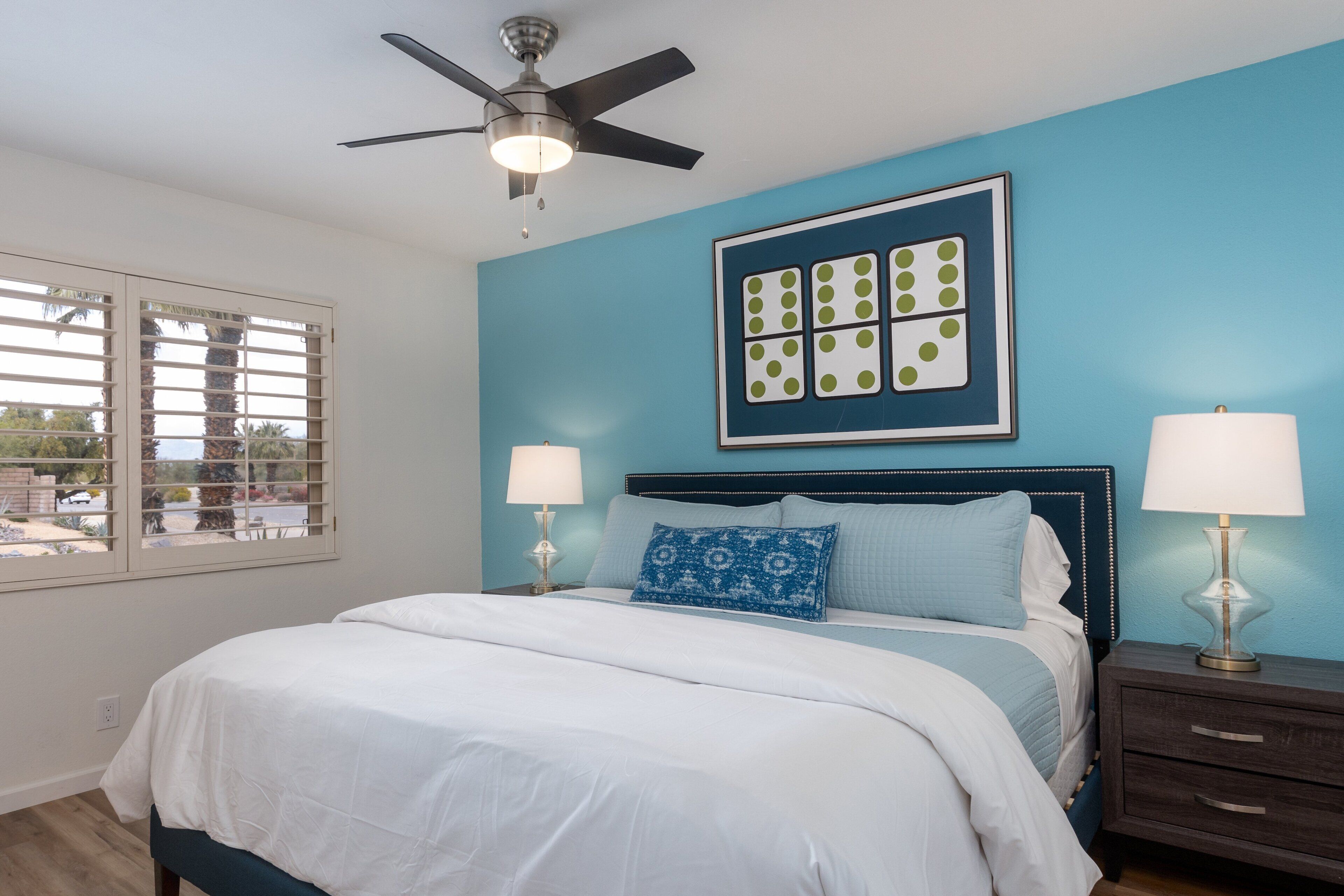 A bedroom with a bed, two bedside tables with lamps, a ceiling fan, and a window with blinds.