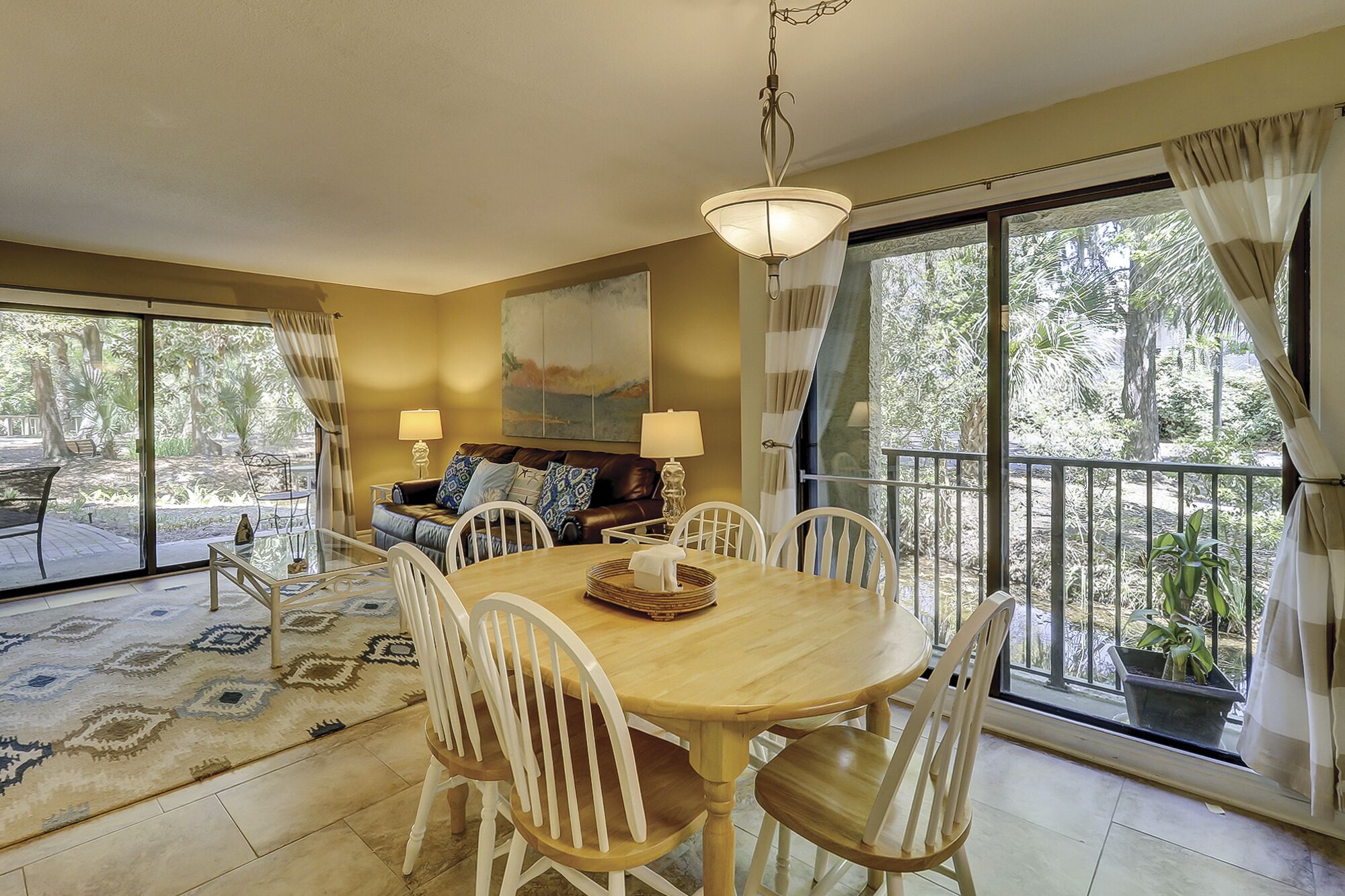 A dining area with a wooden table and white chairs, a sofa with patterned cushions, and a painting above the sofa.