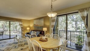 A dining area with a wooden table and white chairs, a sofa with patterned cushions, and a painting above the sofa. - Beach Retreat! Prime Location, Close to Coligny Plaza (Hilton Head Island)