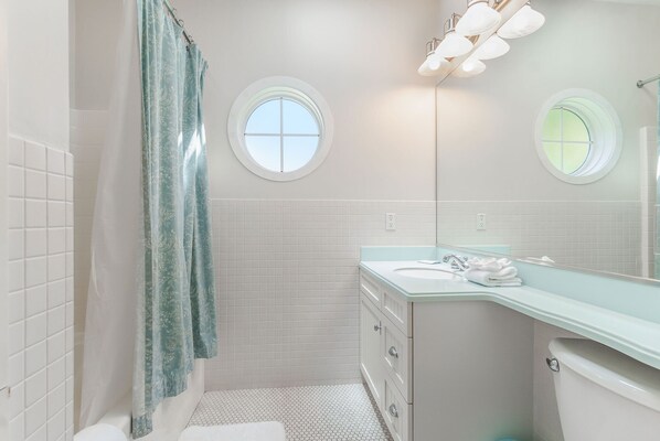 Un baño con paredes de azulejos blancos, un lavamanos blanco y un inodoro. Se ven dos ventanas redondas y una cortina de ducha.