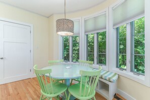 Un coin repas avec une table ronde et des chaises vertes, de grandes fenêtres à cadres blancs et un luminaire suspendu.