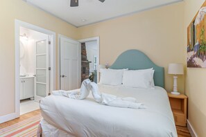 Un lit bien fait, avec une couette blanche et des oreillers, une table de chevet en bois avec une lampe, et un tableau accroché au mur.