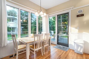 Un espace repas avec une table et des chaises en bois, de grandes fenêtres et une porte donnant sur l’extérieur.