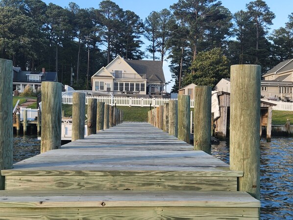 A wooden pier leading to a house with a white fence and tall trees in the background.