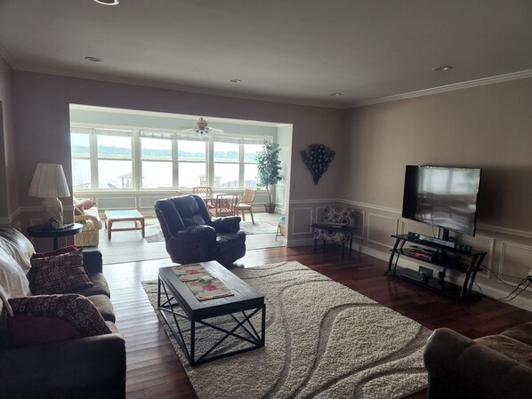 A living room with a sofa, armchair, coffee table, TV, and a view of a body of water through large windows. - Waterfront home with dock, gazebo, and fenced yard (Lewes)