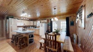 Una cocina de estilo rústico tipo cabaña, con armarios de madera, una isla central con taburetes, una mesa de comedor con sillas y una lámpara colgante.