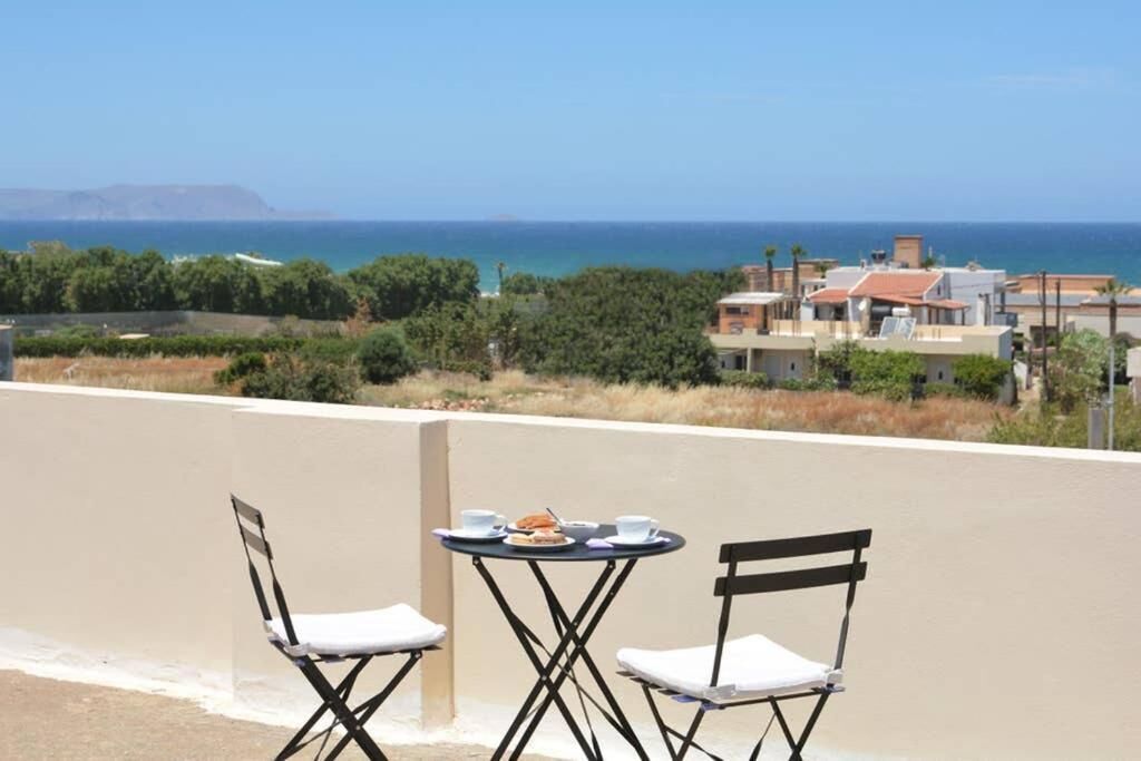 A balcony with two chairs and a small table, overlooking a coastal area with buildings and a clear view of the sea.