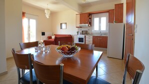 A modern kitchen with a wooden dining table, chairs, and a fruit bowl. There is a refrigerator, oven, and cabinets. - Olive Oil Residence (Hersonissos)