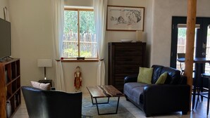 Ein Wohnzimmer mit einem schwarzen Ledersofa, einem Holztisch, einem schwarzen Sessel, einer Holzkommode, einem Fernseher und einem Fenster mit Vorhängen.
