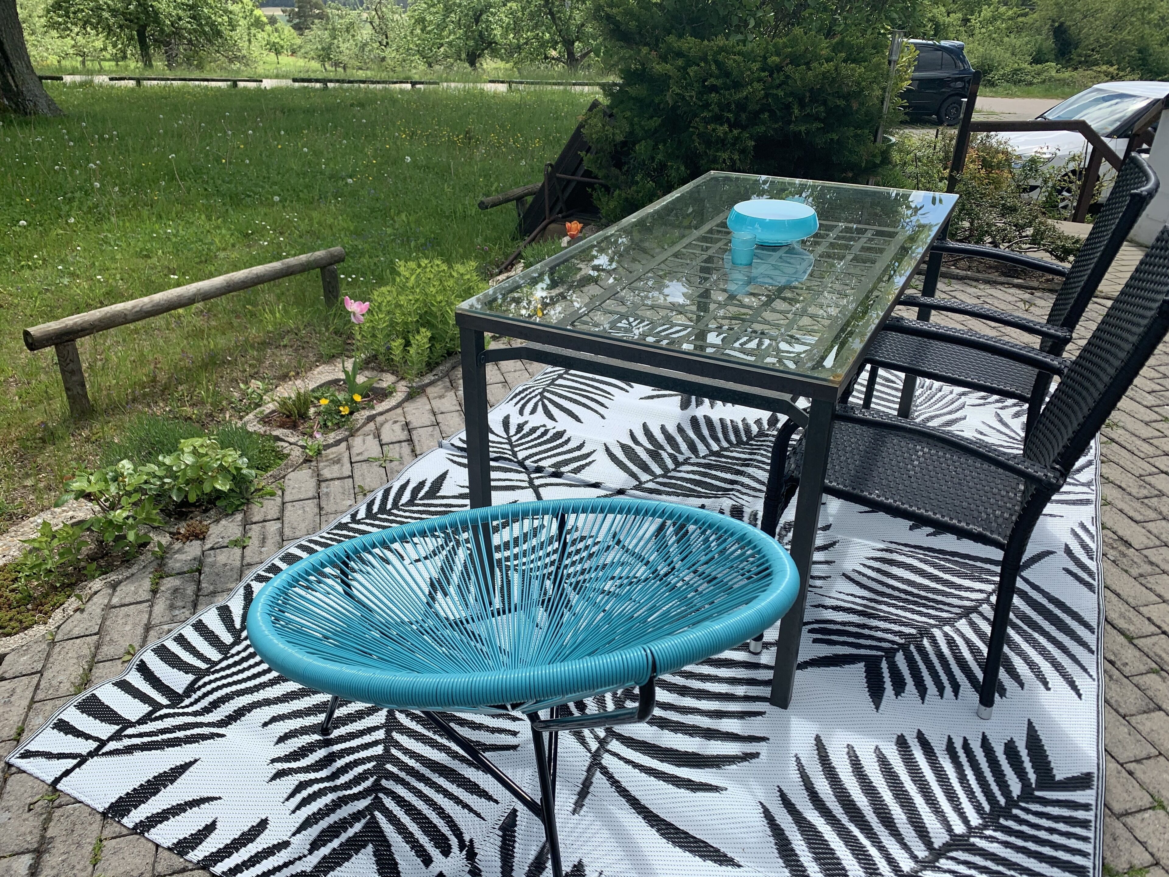 Une table avec un plateau en verre, des chaises et un fauteuil en osier, le tout posé sur un tapis à motifs.