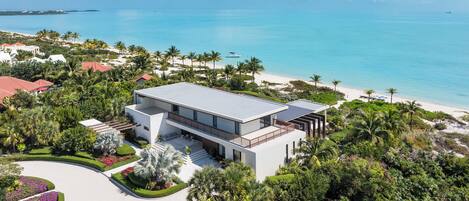 A modern two-story house with a flat roof, surrounded by lush greenery and a beach.