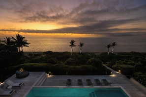 Blick über den Pool mit einem Sonnenuntergang über dem Meer, Palmen und einem wolkenlosen Himmel.