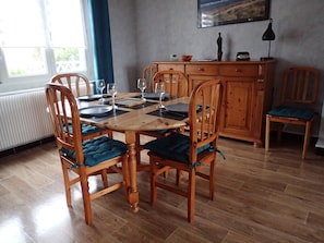 Un coin repas avec une table en bois dressée pour quatre personnes, entourée de chaises en bois dotées de coussins bleus. Un meuble est adossé au mur, et un tableau est accroché au-dessus.