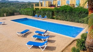 Une piscine rectangulaire à l’eau bleue, entourée d’une terrasse carrelée. Quatre chaises longues bleues sont alignées le long du bord de la piscine. On aperçoit à l’arrière-plan un bâtiment jaune avec des volets bleus et un balcon.