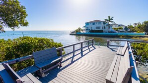 A wooden deck with benches overlooking a body of water, a white building with a balcony, and palm trees in the background. - Peaceful Condo – Cedars East #803 by RVA (Longboat Key)