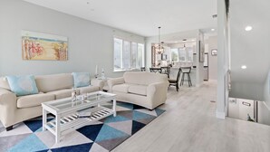 A modern living room with a sofa, coffee table, and a geometric rug. There is a dining area with a table and chairs in the background. - Peaceful Condo – Cedars East #803 by RVA (Longboat Key)