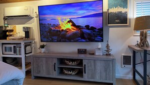 A living room with a flat-screen TV mounted on the wall, a wooden cabinet below, and a framed cityscape picture. - West Seattle Driftwood Cottage - Firepit + Walk to Coffee & Restaurants (Seattle)