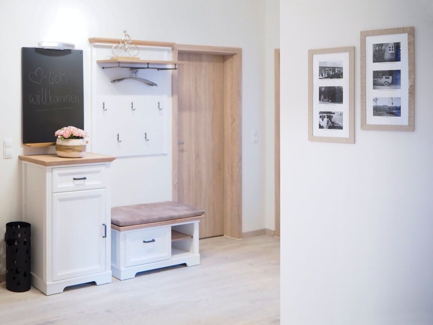 A small, neatly organized room with a white cabinet, a bench, a chalkboard with a welcome message, a coat rack, and framed photographs on the wall.
