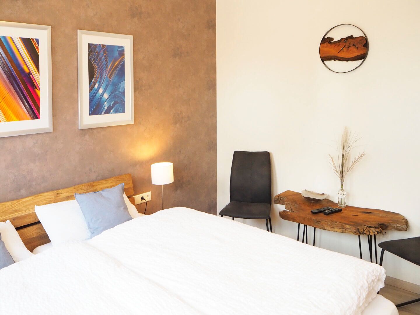 A hotel room with a bed, a desk, a chair, and two framed artworks on the wall.