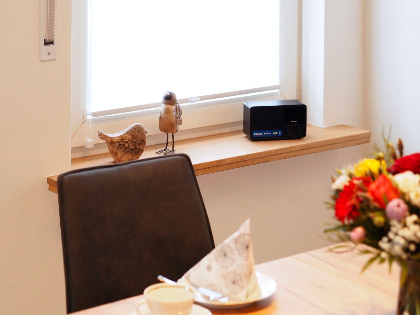 Zona pranzo con tavolo in legno, una sedia, un piccolo uccellino decorativo, un altoparlante Bluetooth e una finestra con luce naturale.