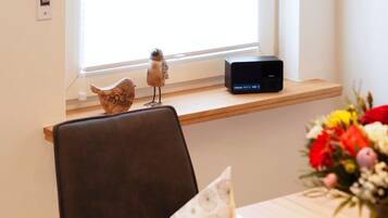 Zona pranzo con tavolo in legno, una sedia, un piccolo uccellino decorativo, un altoparlante Bluetooth e una finestra con luce naturale.