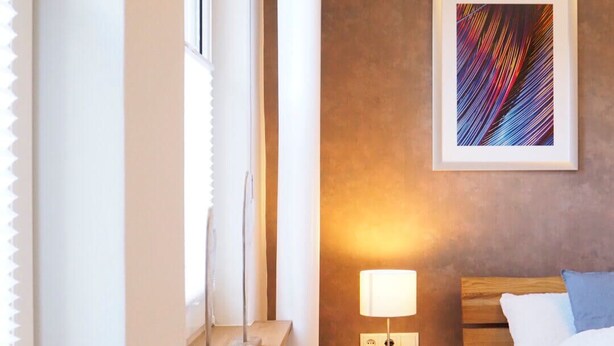 Una habitación de hotel moderna con una cama de madera, una mesita de noche con lámpara, un cuadro en la pared y una planta en maceta.