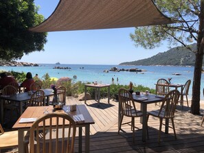 Un espace de restauration en plein air, avec des tables et des chaises en bois, offrant une vue sur la côte, avec une eau d’un bleu limpide et des affleurements rocheux.