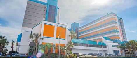 Un hotel con un diseño arquitectónico moderno, que destaca por una llamativa combinación de colores azul y naranja, un amplio estacionamiento y palmeras al frente.