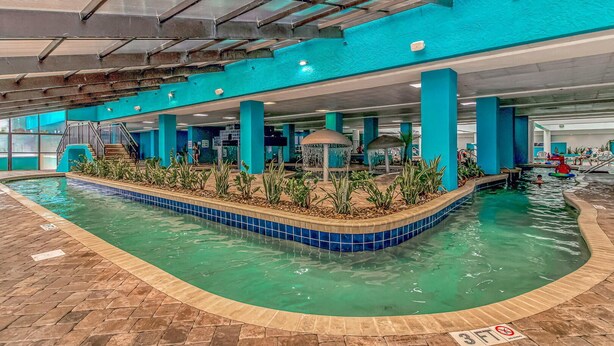 An indoor swimming pool with a tiled edge, a central island with plants, and a covered seating area. - Landmark 607 (Myrtle Beach)