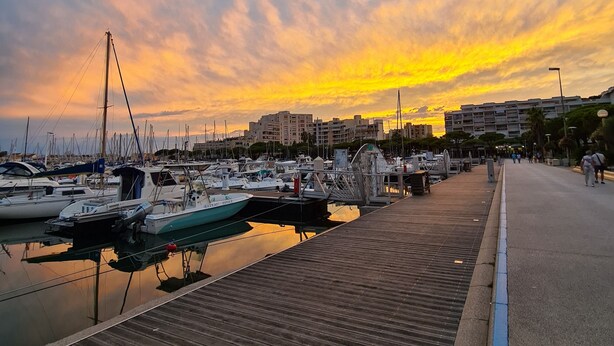 Un puerto deportivo con barcos amarrados, un muelle de madera y edificios al fondo.