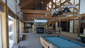 A pool table in a room with a stone fireplace, wooden beams, and large windows.