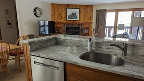 A kitchen with a marble countertop, stainless steel sink, wooden cabinets, a dishwasher, a television, and a fireplace.