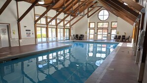 An indoor swimming pool with a high ceiling and wooden beams, surrounded by windows and a seating area.