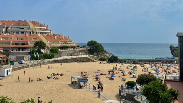 Una playa con gente tomando el sol y caminando, una torre de salvavidas y edificios al fondo.