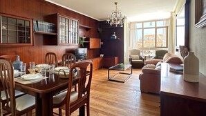 Amplia sala de estar con una mesa de comedor de madera para cuatro personas, un sofá cómodo y un gran ventanal con vistas a edificios.