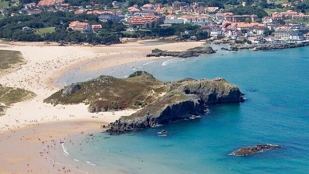 An aerial view of a coastal area with a beach, rocky outcrops, and a town in the background.