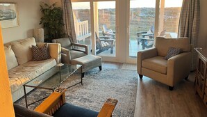 A spacious living room with a sofa, armchairs, a glass coffee table, and a view through large sliding glass doors to an outdoor deck with chairs and a table. - Amelia Island oceanfront home on Sandpiper Loop, amazing views & steps to ocean (Fernandina Beach)