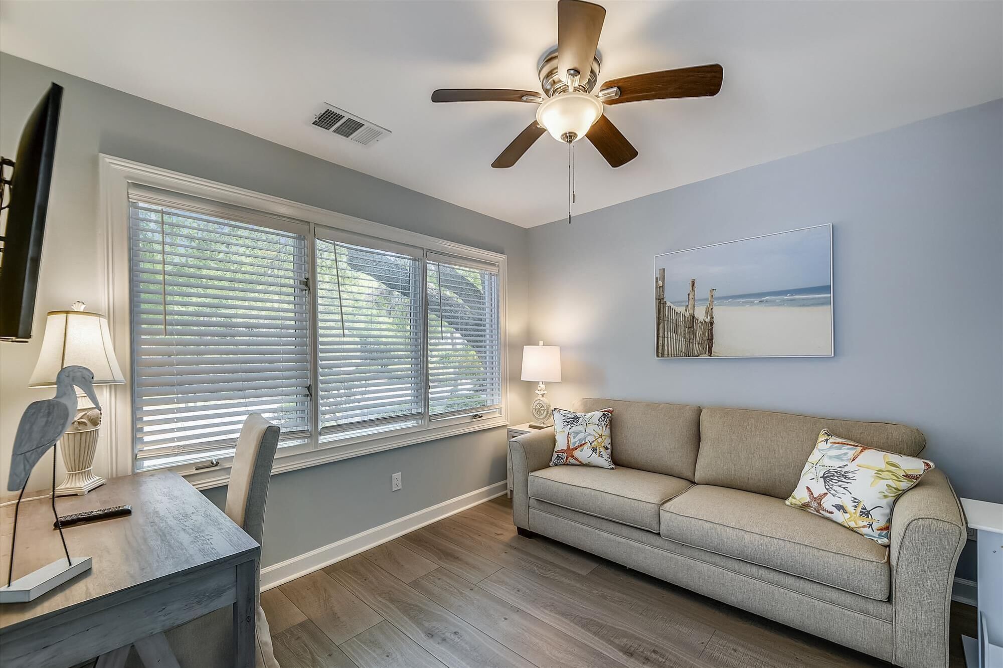 A living room with a ceiling fan, a sofa with floral cushions, a table with a lamp, and a beach-themed painting on the wall.