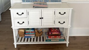 A white wooden console table with drawers and a shelf, decorated with a framed painting of the ocean, a welcome sign, and several books. - 4 BR beach house 2 blocks from ocean in Cherry Grove! Dog friendly! (North Myrtle Beach)