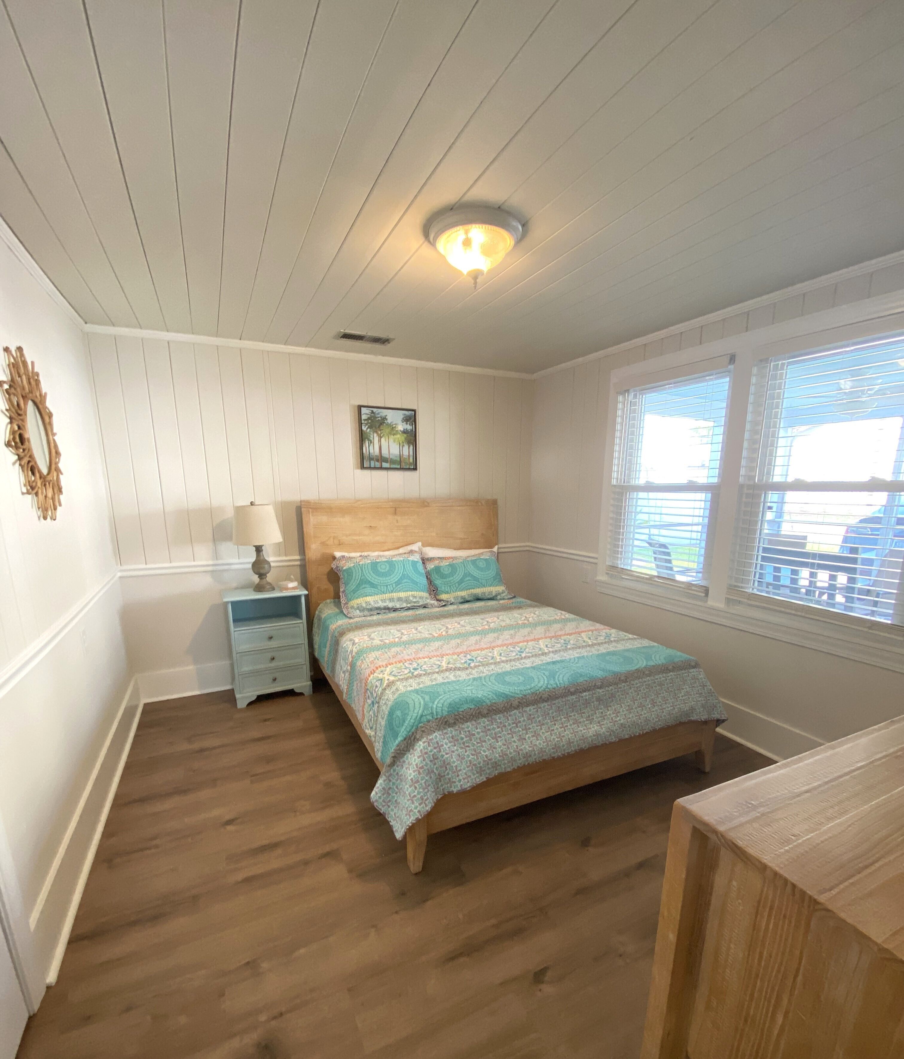 Un dormitorio con cama de madera, una mesita de noche, una lámpara, una luz de techo, una ventana con persianas y un espejo montado en la pared.