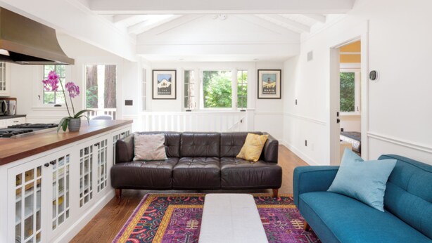 Een ruime woonkamer met een donker leren bank, een blauwe fauteuil en een kleurrijk vloerkleed.