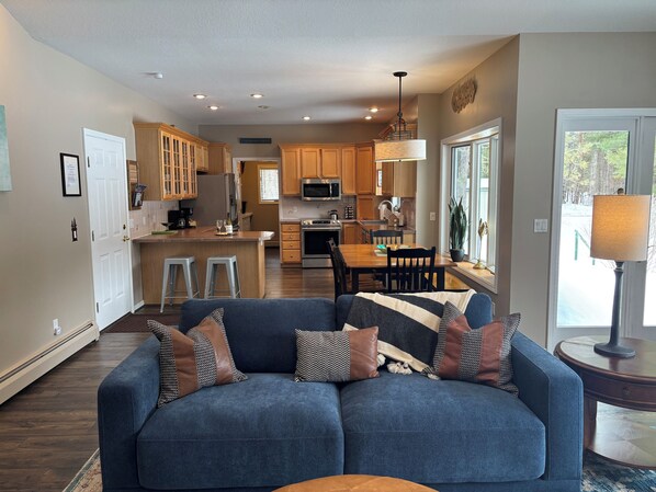 A modern kitchen with wooden cabinets, a dining area with a table and chairs, and a living room with a sofa and a lamp.