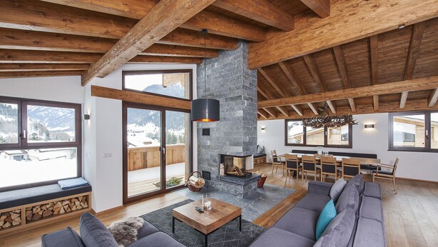 Amplia sala de estar con chimenea de piedra, vigas de madera, un sofá grande y un comedor con vistas a las montañas.
