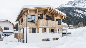 Una casa moderna de varios pisos de madera, con un balcón, rodeada de nieve y montañas al fondo.