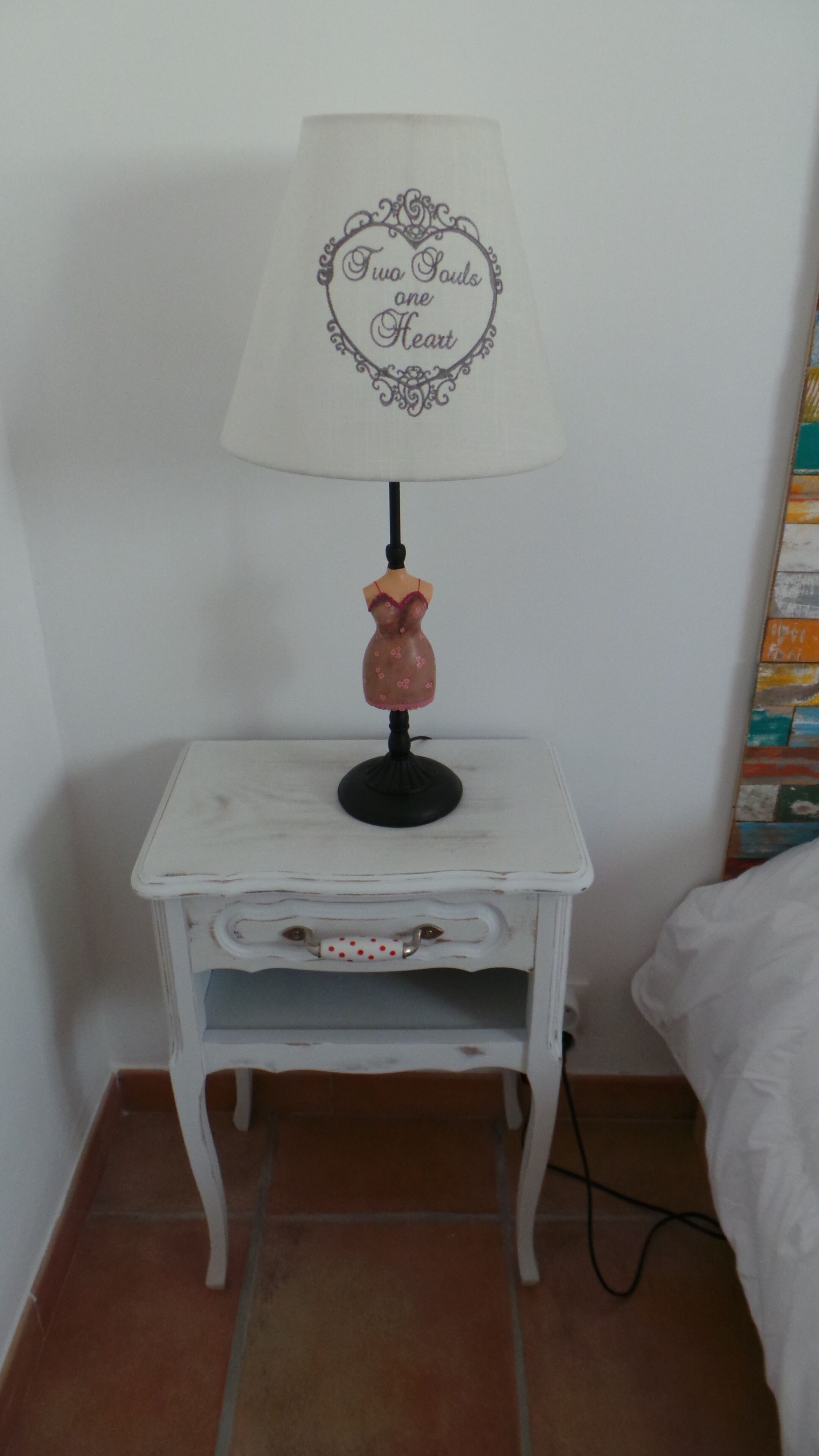 Een klein nachtkastje, wit, met een lamp met een decoratieve lampenkap en een voet in vintage stijl.